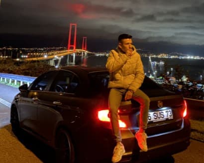 Man in beige jacket sits on car trunk overlooking a red-lit bridge at night.