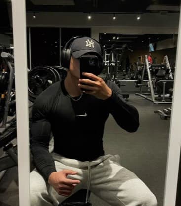 Man in black Nike shirt, grey sweatpants, and Yankees cap taking a gym mirror selfie.